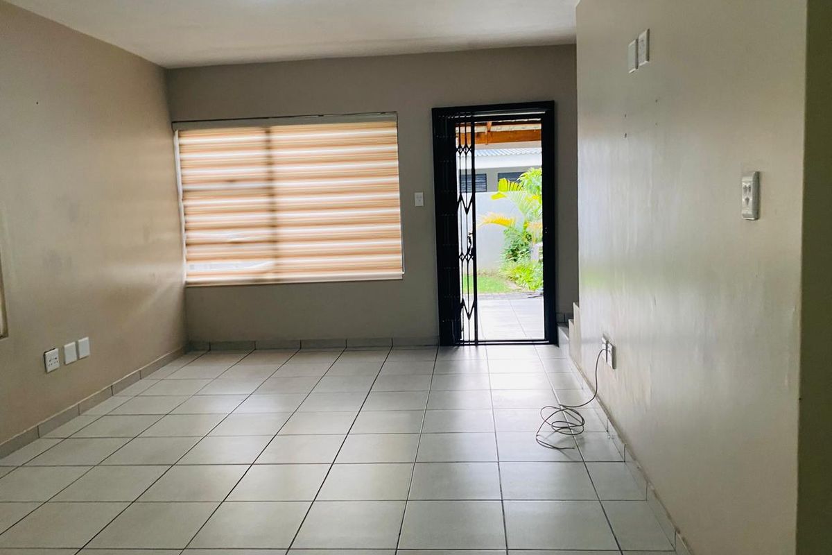A bright, open-plan living area featuring neutral tiled flooring, a large window with fitted blinds, and direct access to an outdoor patio area.