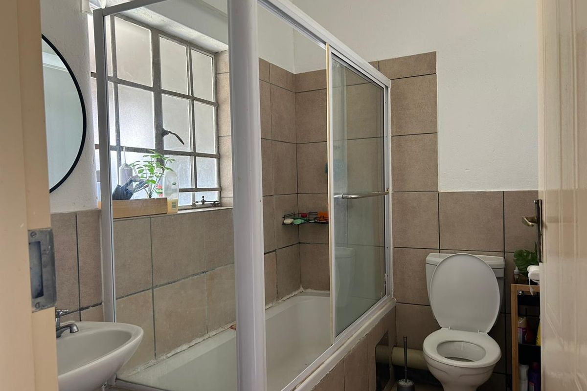 A functional bathroom featuring a shower-over-bath combination, a pedestal basin, and a toilet, finished with neutral wall tiling and a steel-framed window.