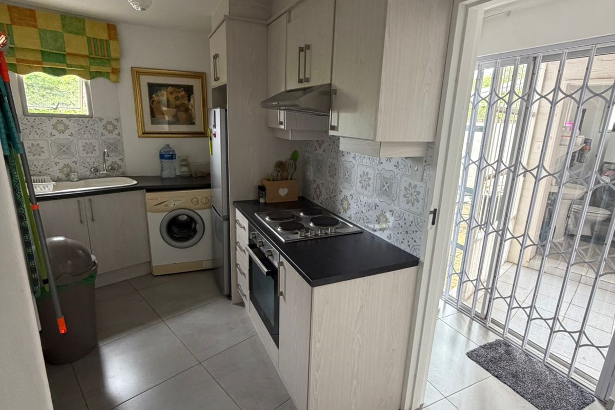 Compact kitchen featuring light wood-grain cabinetry, dark countertops, built-in oven, hob, extractor fan, and tiled flooring with direct access to an outdoor area.