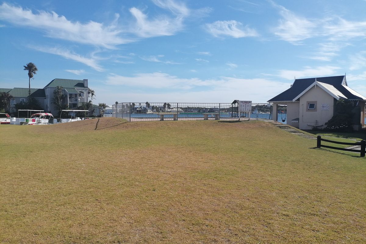 A well-maintained communal lawn area featuring a fenced perimeter, overlooking the water with adjacent residential buildings and a small utility structure.