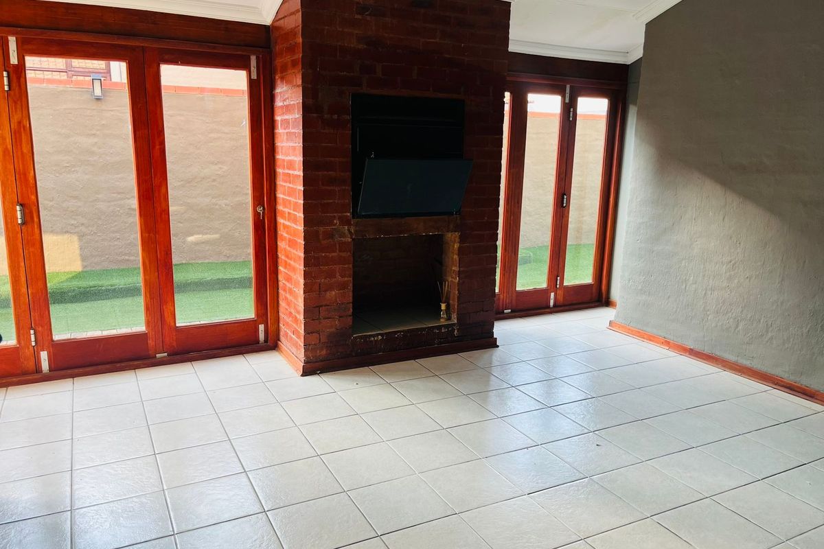 An empty living room featuring an exposed brick fireplace, tiled flooring, and large wooden-framed glass doors providing access to the outdoor area.