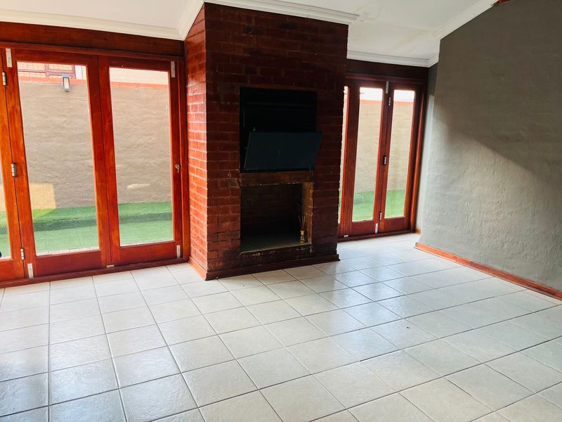 An empty living room featuring an exposed brick fireplace, tiled flooring, and large wooden-framed glass doors providing access to the outdoor area.