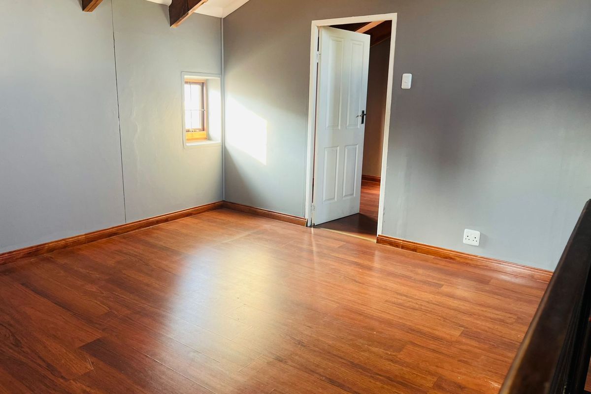An empty bedroom featuring warm laminate flooring, exposed timber ceiling beams, and a small window, providing a clean and neutral space.