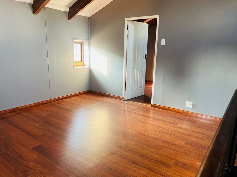 An empty bedroom featuring warm laminate flooring, exposed timber ceiling beams, and a small window, providing a clean and neutral space.