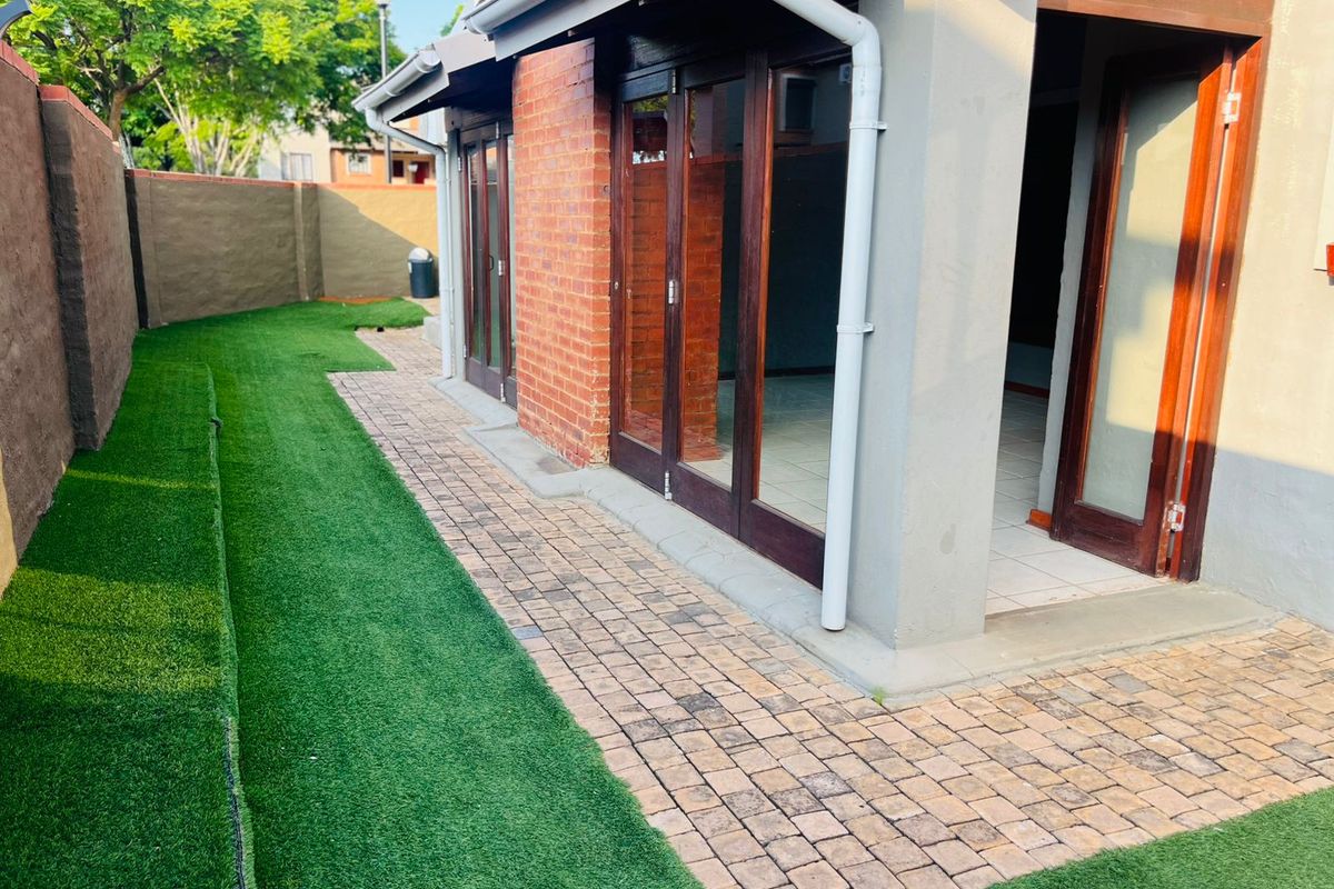 Rear exterior view of the townhouse featuring a paved patio area, artificial lawn, and timber-framed glass doors leading into the living space.
