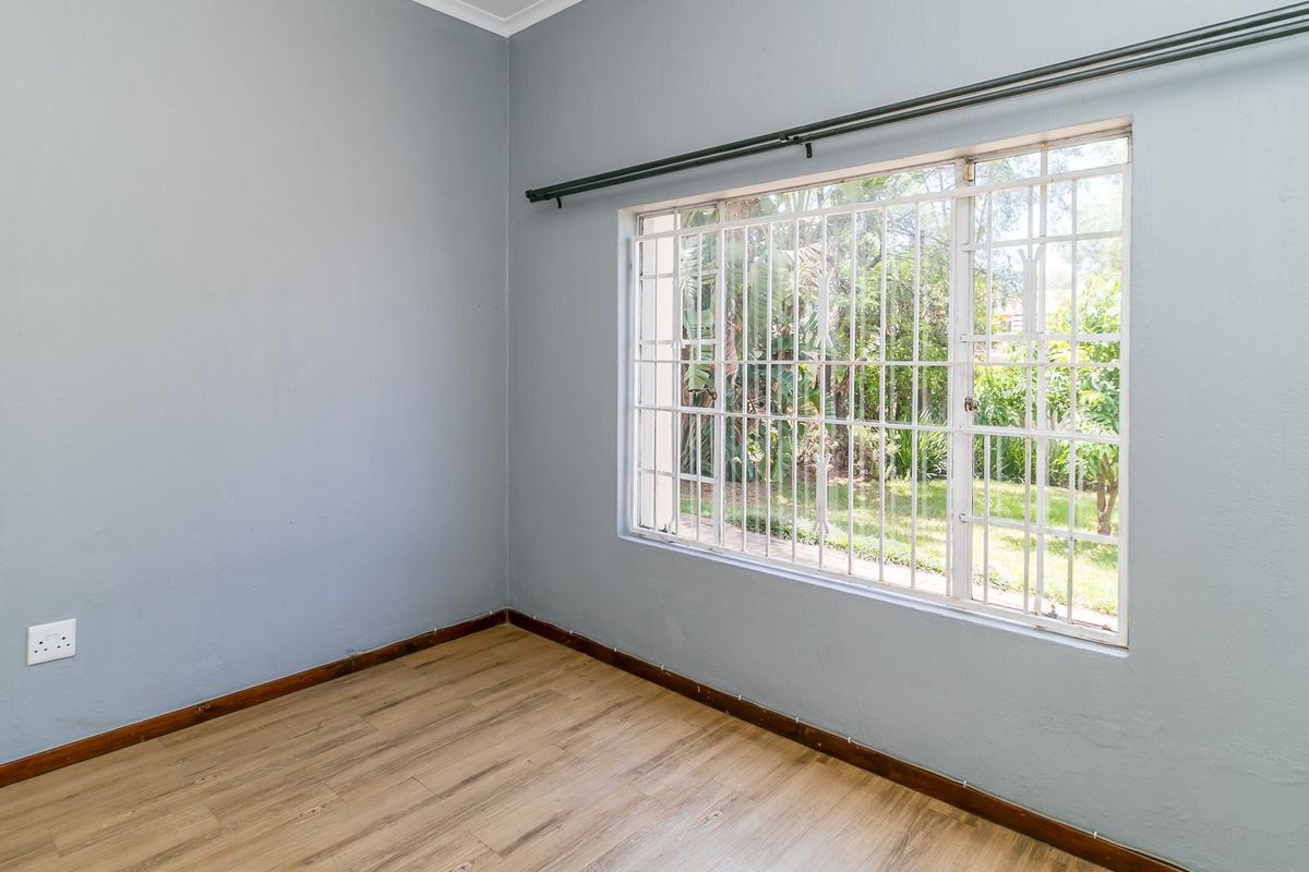 A bright, empty bedroom featuring laminate flooring, neutral grey walls, and a window fitted with security bars and a curtain rail.