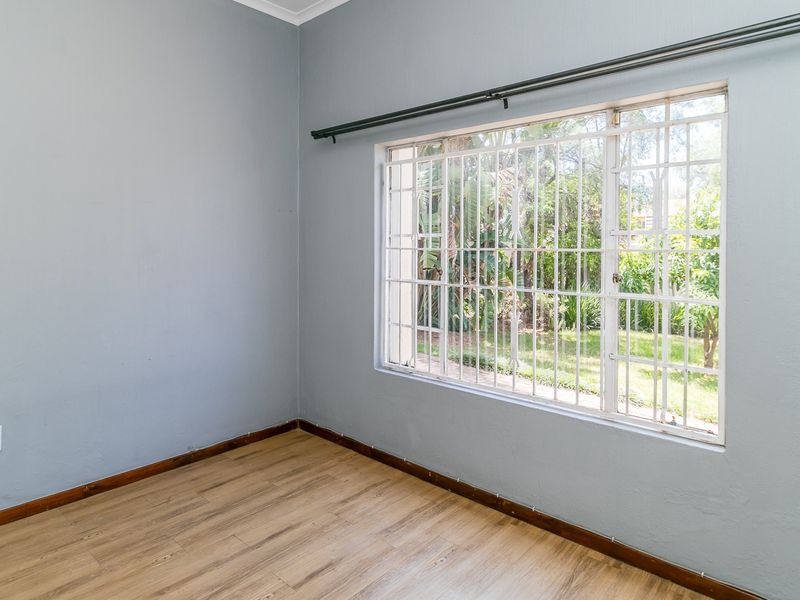 A bright, empty bedroom featuring laminate flooring, neutral grey walls, and a window fitted with security bars and a curtain rail.