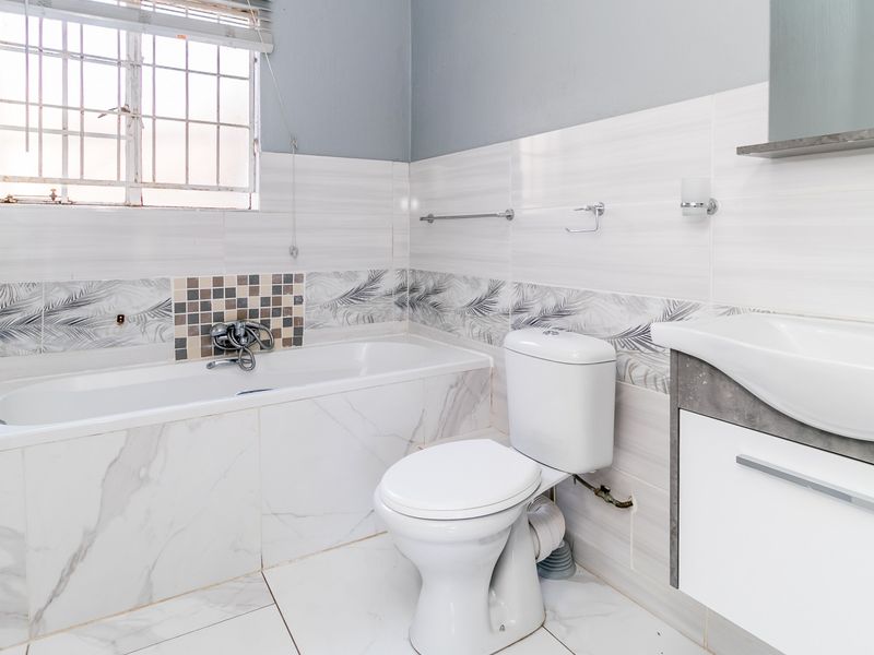 A clean, functional bathroom featuring a bathtub, toilet, and vanity basin with modern tiled finishes and a window with security bars.