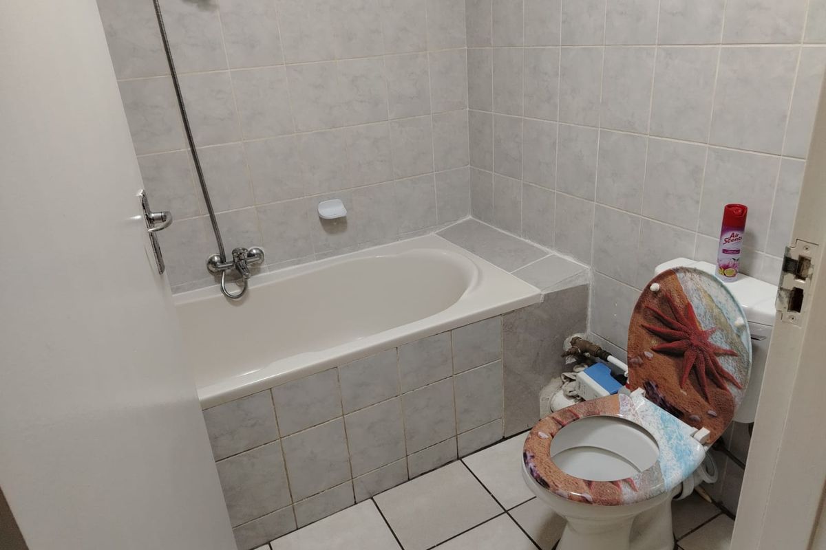 A compact bathroom featuring a built-in bathtub with shower mixer and tiled finishes, requiring minor cosmetic updates to the exposed plumbing.