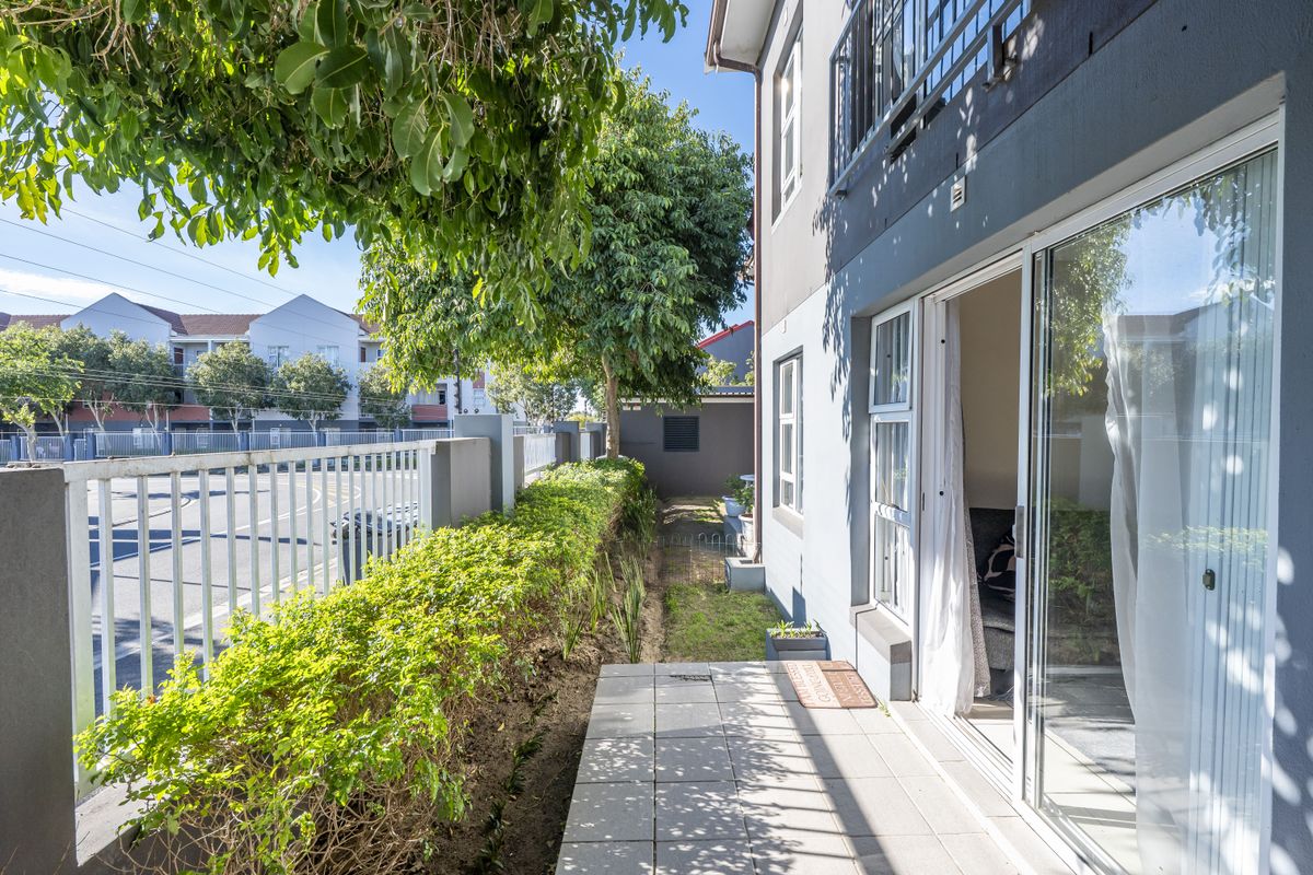 A paved rear patio area featuring sliding glass door access, bordered by a manicured hedge and metal perimeter fencing.