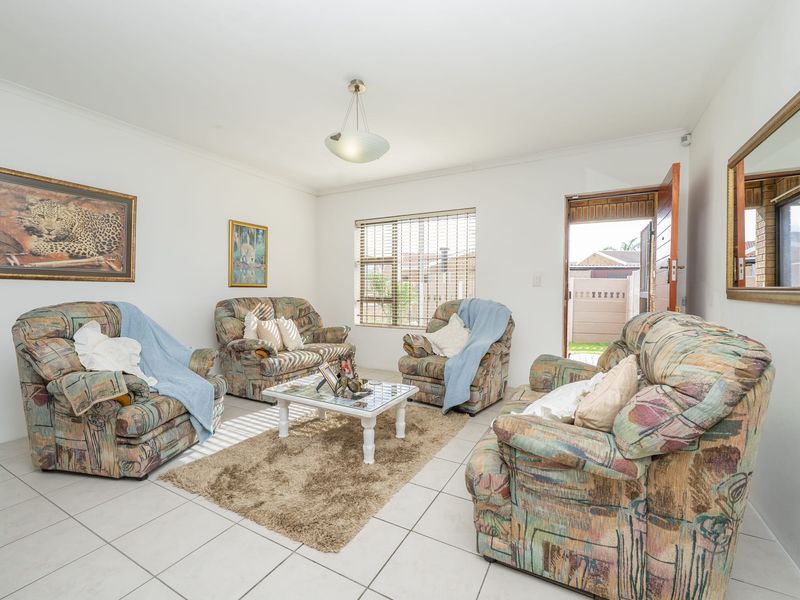 A bright living room featuring neutral tiled flooring, a central pendant light, and natural light through venetian blinds with direct access to the exterior.
