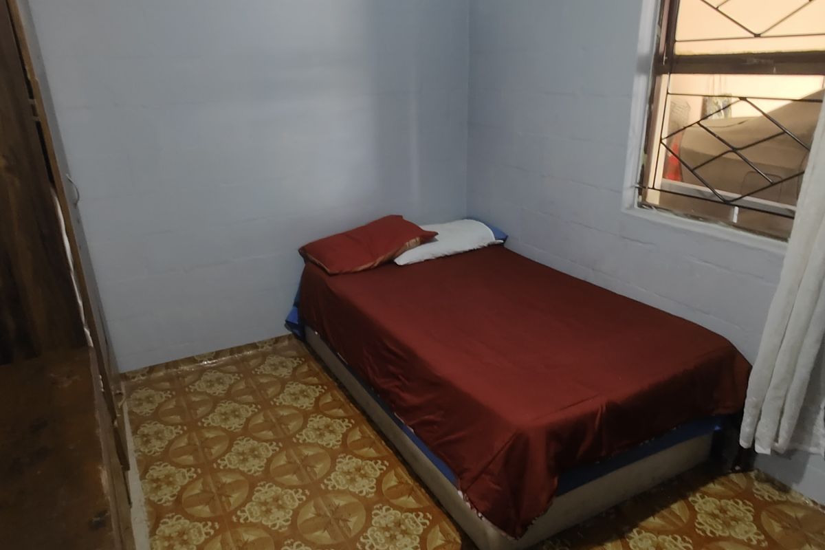 A simple bedroom featuring exposed blockwork walls, patterned floor tiles, and a window fitted with security bars.