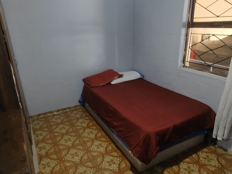 A simple bedroom featuring exposed blockwork walls, patterned floor tiles, and a window fitted with security bars.