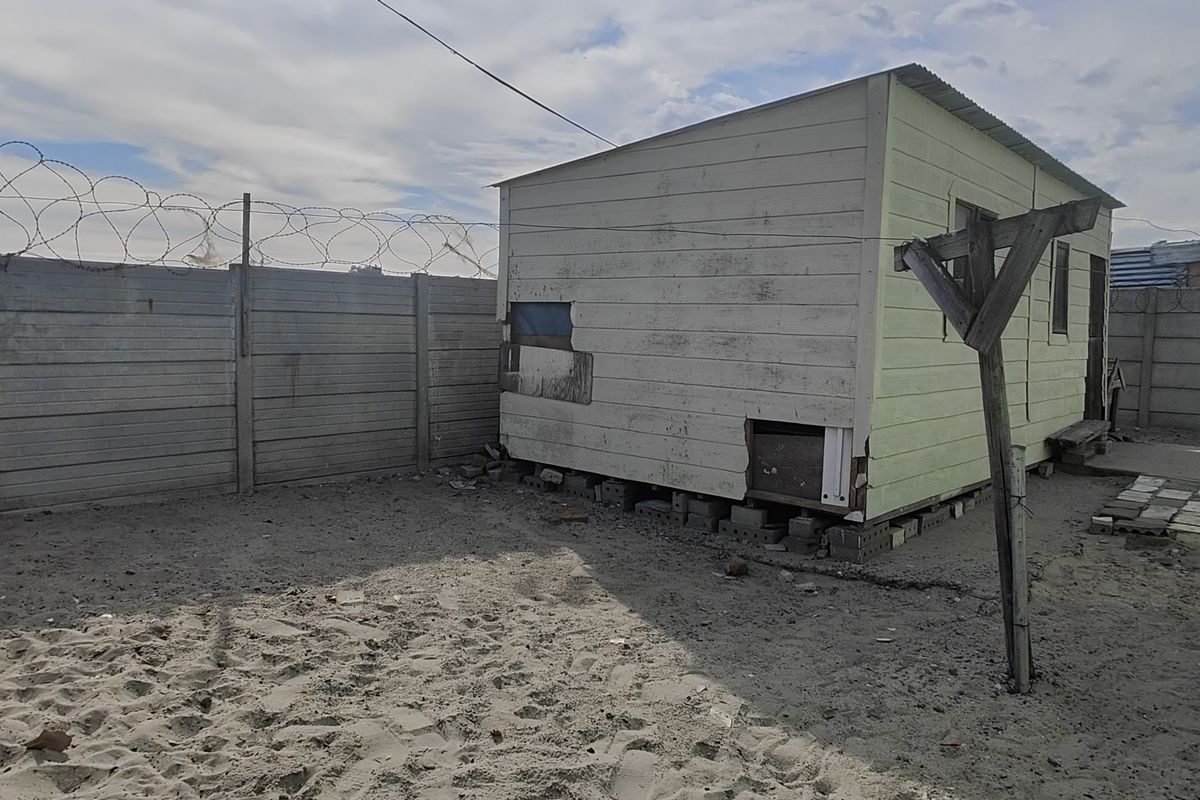 Rear view of a timber-clad structure featuring a precast concrete boundary wall with barbed wire security topping and a sandy yard area.