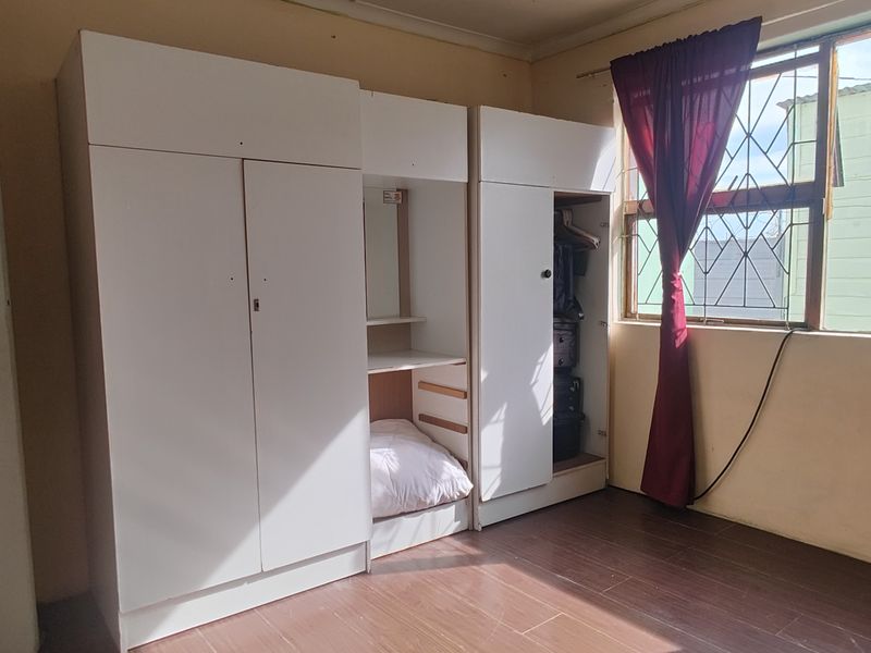 A bedroom featuring built-in white cupboards, laminate flooring, and a window fitted with security bars.