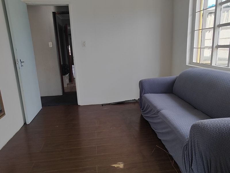 A living room area featuring dark laminate flooring and a window with security bars, showing signs of wear on the floor and skirting.