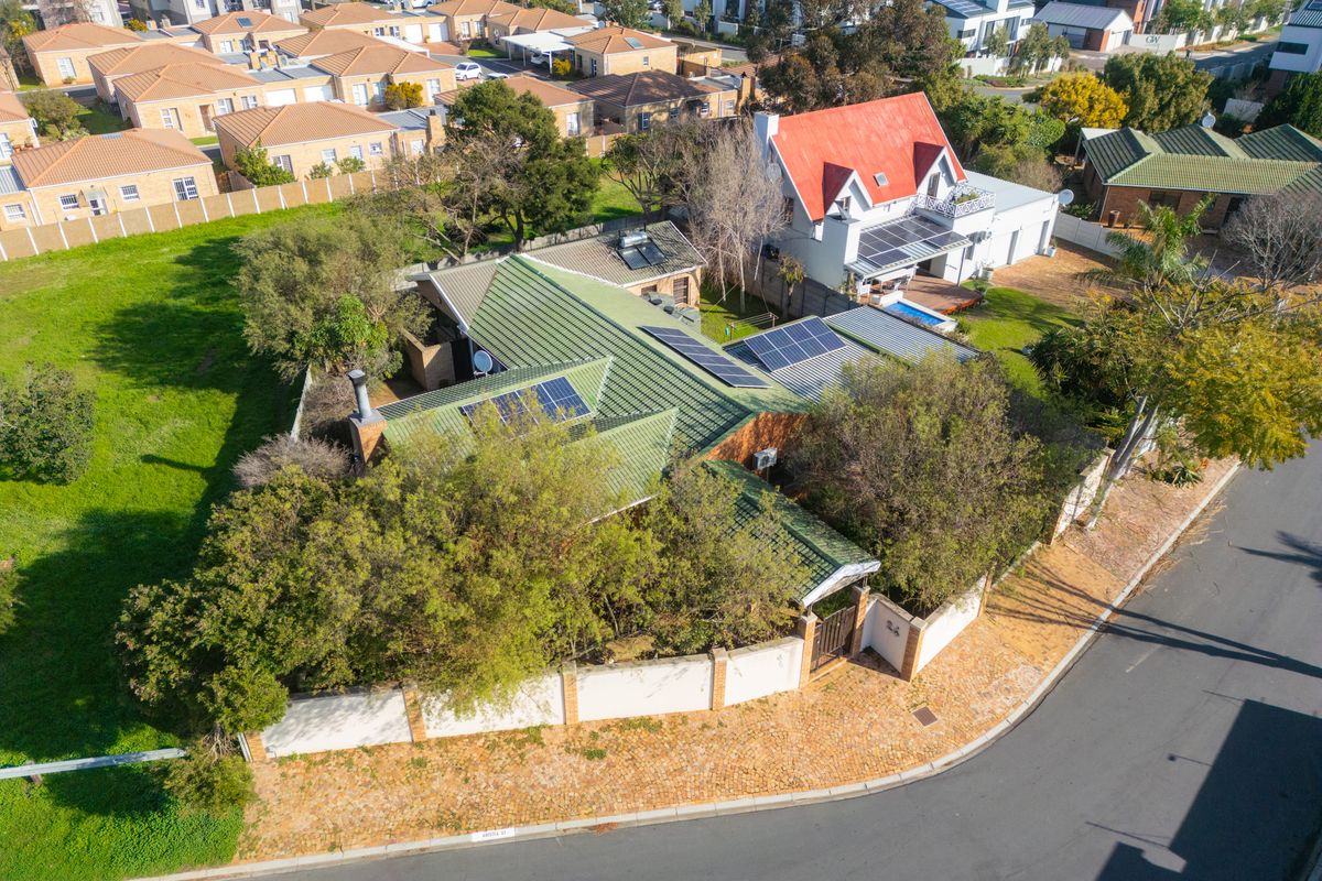 Aerial view of a residential property featuring a green metal roof, installed solar panels, and a secure perimeter wall in a suburban neighborhood.