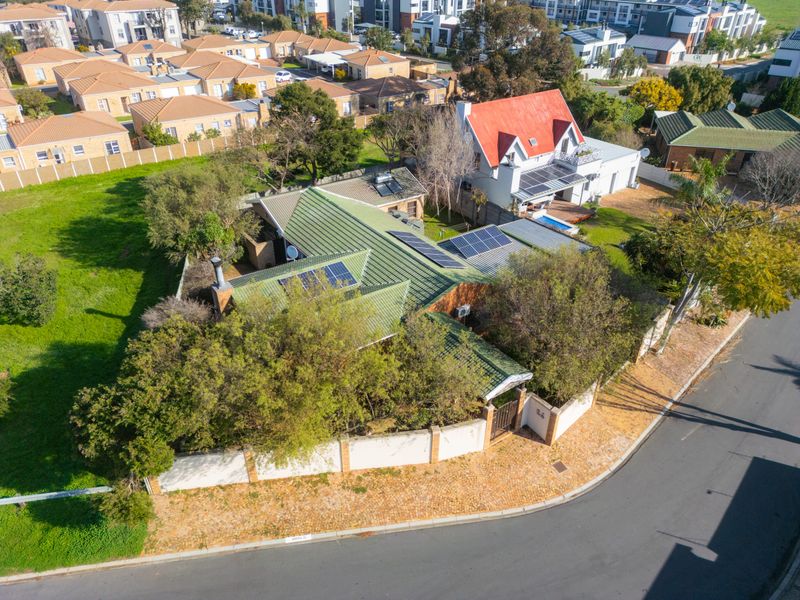 Aerial view of a residential property featuring a green metal roof, installed solar panels, and a secure perimeter wall in a suburban neighborhood.
