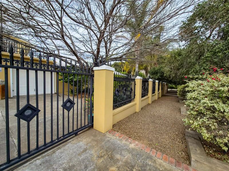Front exterior view featuring a secure wrought iron gate, masonry boundary wall, concrete driveway, and a gravel pathway leading along the property perimeter.