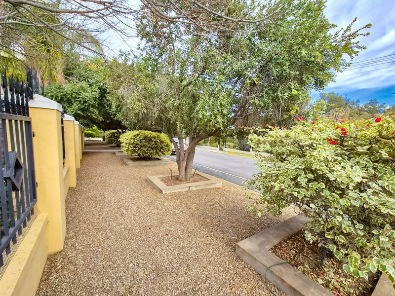 A street-level view of the property boundary featuring a secure wrought iron fence, concrete pillars, and established garden beds with gravel landscaping.