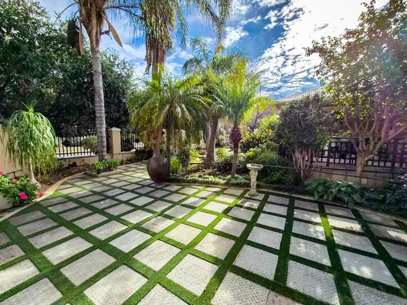A well-maintained garden area featuring a grid-patterned paved patio, mature palm trees, and established landscaping with perimeter fencing.