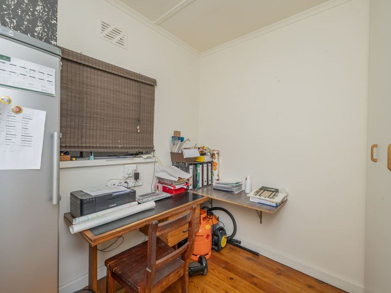 A compact home office space featuring timber flooring, built-in storage cabinetry, and a wall-mounted desk area with a window blind.