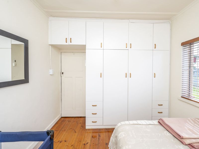 A bedroom featuring polished timber flooring, extensive white built-in cabinetry with overhead storage, and a window fitted with venetian blinds.