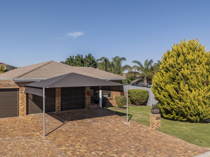 Front view of a brick house featuring a double garage, paved driveway, and a modern carport structure set within a well-maintained garden.