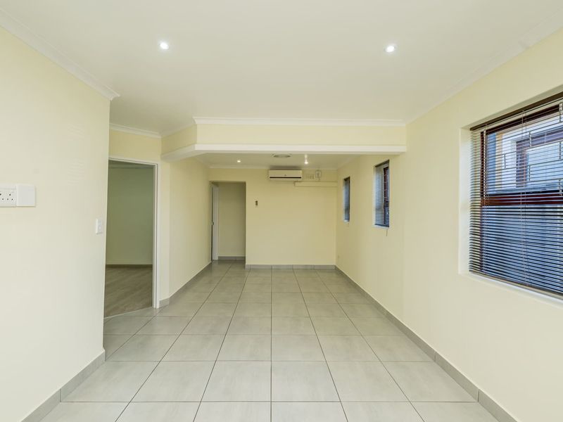 A bright, empty living area featuring neutral tiled flooring, recessed downlights, an air conditioning unit, and venetian blinds on the windows.