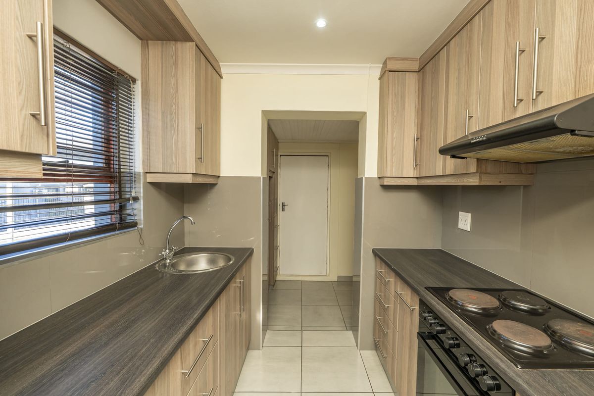 A clean, modern galley-style kitchen featuring wood-look cabinetry, dark laminate countertops, integrated electric cooking appliances, and neutral tiled finishes.