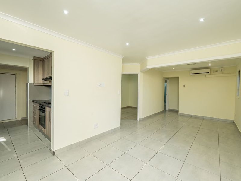 An open-plan living area featuring neutral tiled flooring, recessed lighting, and a split-system air conditioner, seamlessly connected to a compact kitchen space.