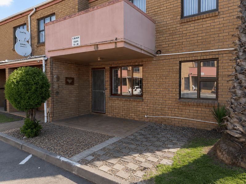 Front exterior view of a brick apartment block featuring a paved entrance, security gate, and an overhead balcony.