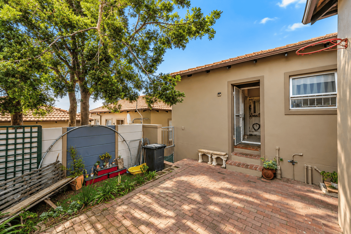 A rear courtyard area featuring brick paving, a security gate entrance, and a boundary wall with established garden elements.