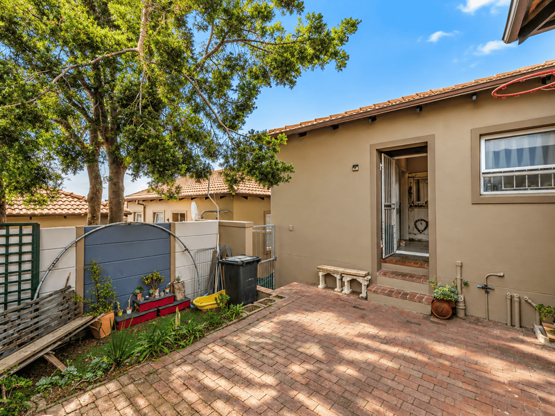 A rear courtyard area featuring brick paving, a security gate entrance, and a boundary wall with established garden elements.