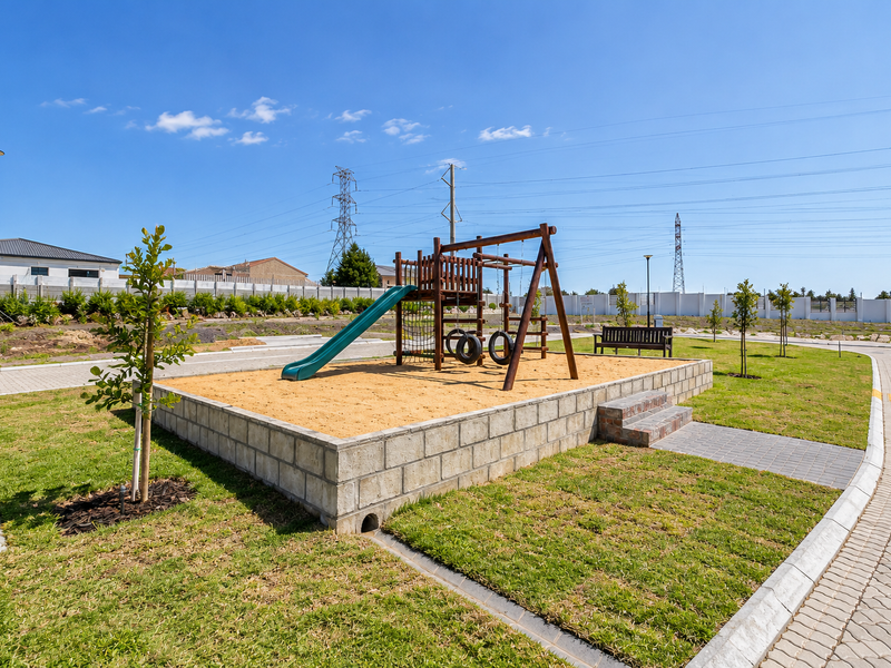 A well-maintained communal garden area featuring a raised playground structure with a slide and swings, set within a landscaped lawn and paved surroundings.