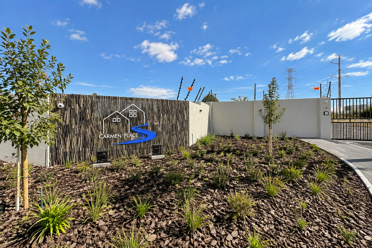 The entrance to Carmen Place featuring a modern stone-clad signage wall, landscaped garden beds, and secure perimeter fencing with an automated gate.