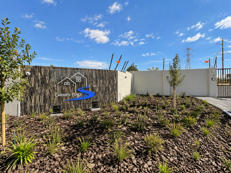 The entrance to Carmen Place featuring a modern stone-clad signage wall, landscaped garden beds, and secure perimeter fencing with an automated gate.
