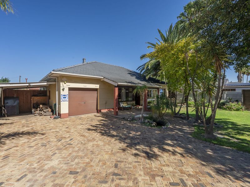 Front view of a single-story house featuring a brick-paved driveway, a roller garage door, and a covered patio area set within a landscaped garden.