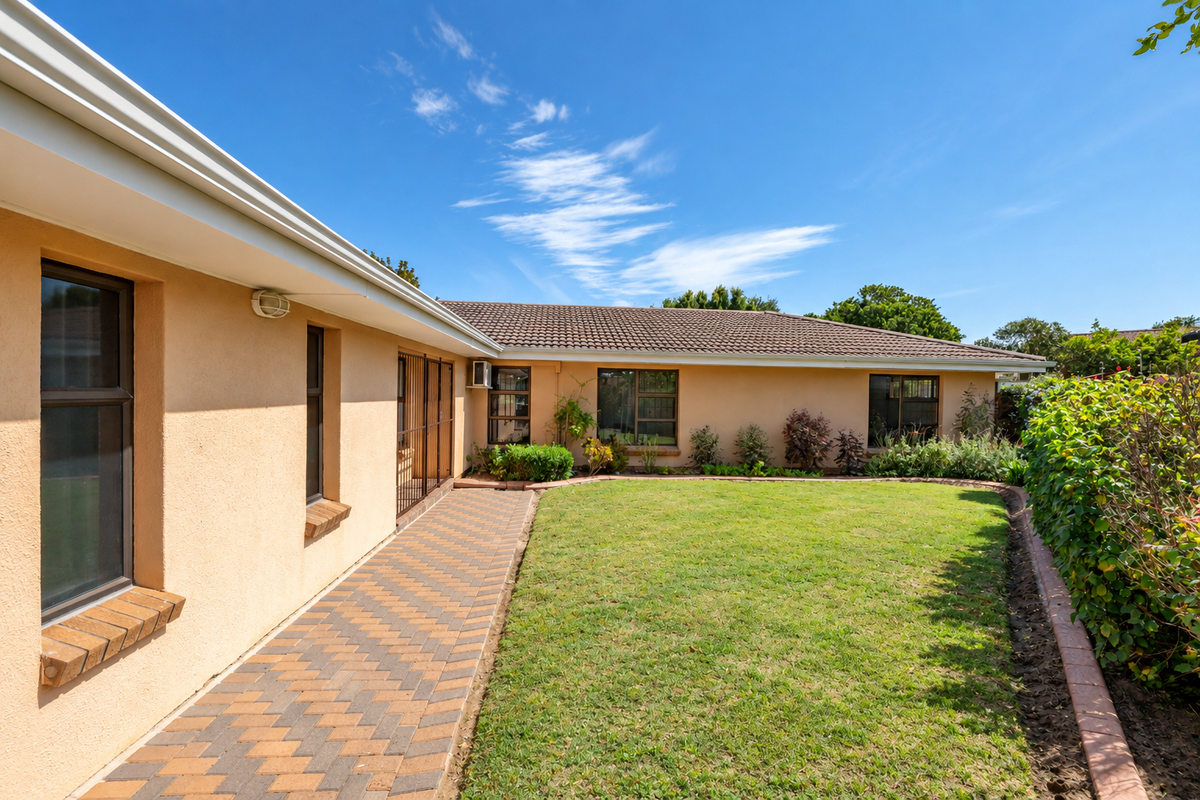 A well-maintained rear exterior view featuring a neat lawn, a brick-paved walkway, and established garden borders against the house facade.