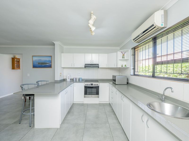 A bright, modern kitchen featuring white cabinetry, stone countertops, integrated appliances, and tiled flooring, with an adjacent breakfast bar and air conditioning unit.