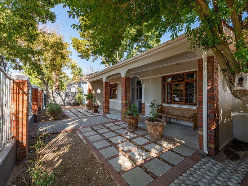 Front elevation of a charming house featuring a covered veranda, brick pillars, and a paved entrance path surrounded by mature trees and garden.