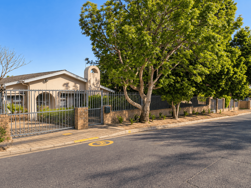 Front exterior view of the property featuring a paved driveway, secure metal fencing, and a brick boundary wall with mature trees.