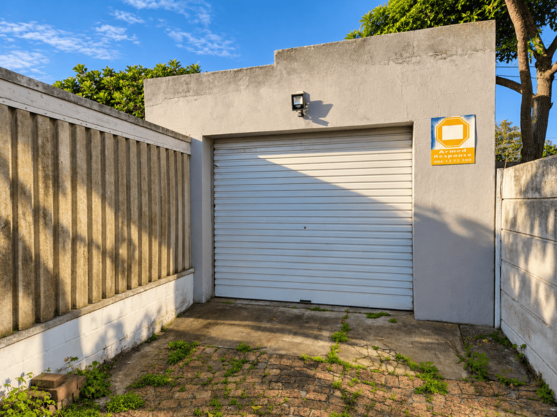 A single garage featuring a white roller shutter door, security lighting, and a paved driveway area with visible weathering and maintenance requirements.