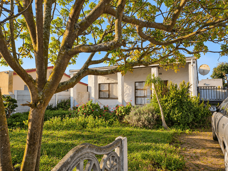 Front view of a single-story house featuring a paved driveway, established garden, and white exterior walls with aluminum window frames.