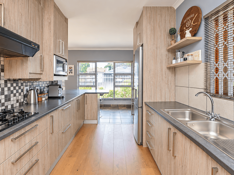 Modern galley-style kitchen featuring light wood-grain cabinetry, dark countertops, gas hob, integrated microwave, and a double stainless steel sink with a window view.