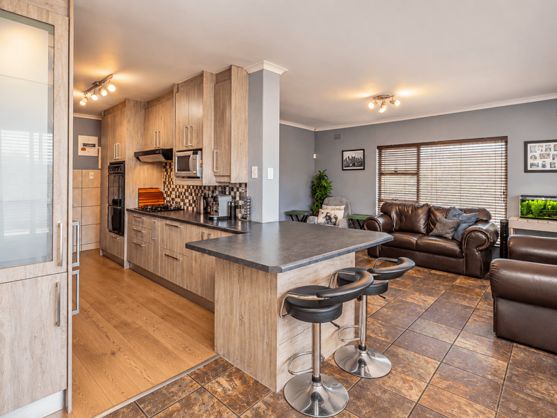 Open-plan kitchen and living area featuring wood-look cabinetry, a breakfast bar, integrated appliances, and a combination of tiled and laminate flooring.