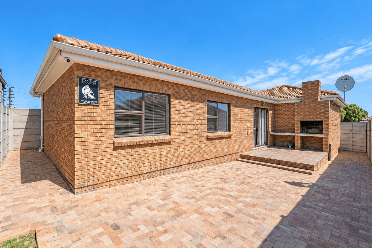Rear exterior view featuring a paved patio area, built-in brick braai, and low-maintenance face-brick facade with aluminium window frames.