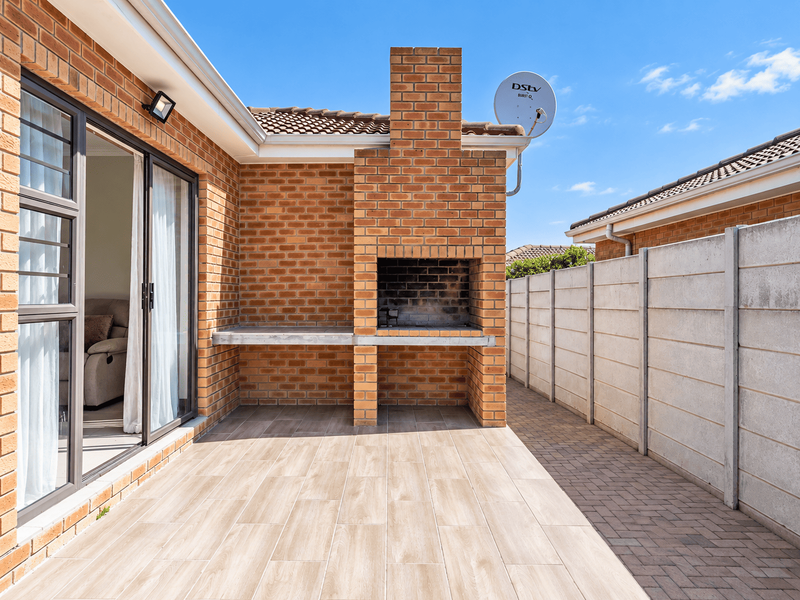 A well-maintained outdoor patio featuring a built-in brick braai, wood-look tiled flooring, and direct access to the interior through sliding glass doors.