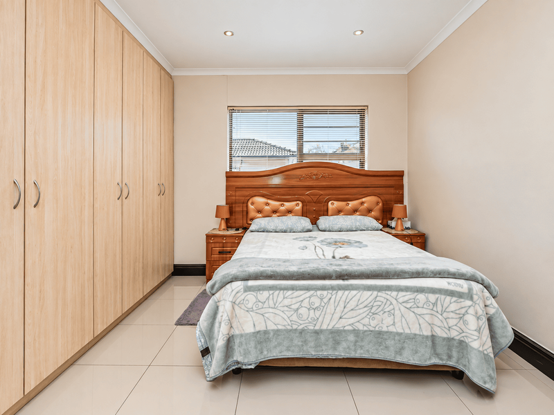 A well-maintained bedroom featuring extensive built-in wooden cupboards, neutral tiled flooring, and recessed ceiling lighting, providing a clean and functional living space.