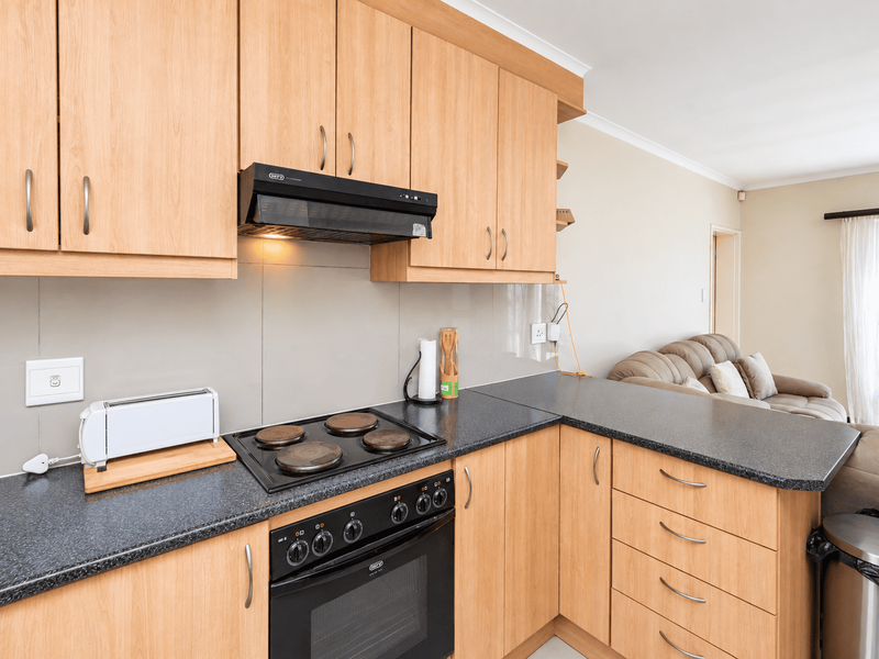 Open plan kitchen featuring light wood cabinetry, dark laminate countertops, built-in oven, and electric hob, seamlessly flowing into the adjacent living area.